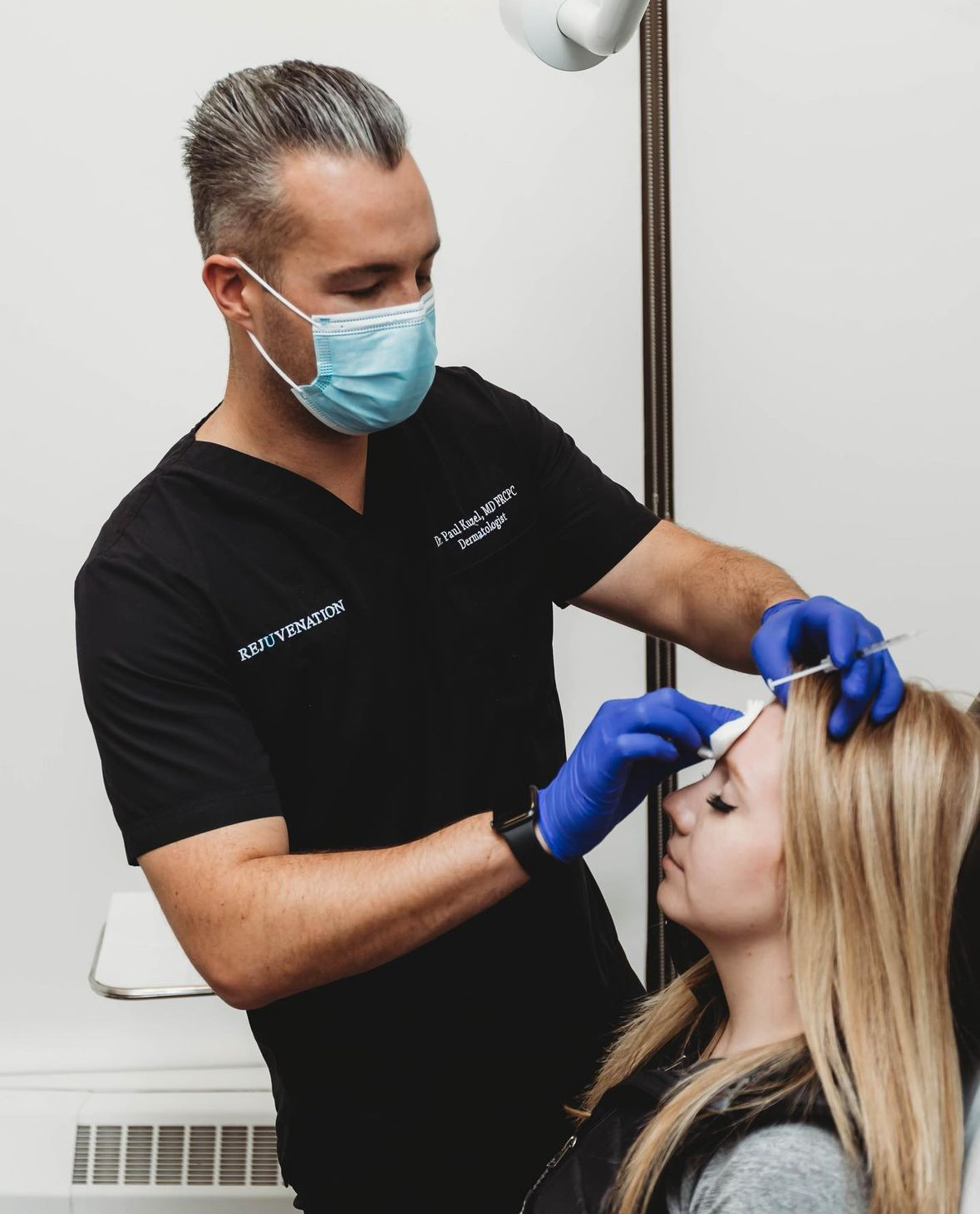 A doctor wearing a face mask and blue gloves is performing a cosmetic Botox® procedure on a female patient. The doctor is holding a syringe and applying a cotton pad to the patient's forehead, indicating the administration of an injectable treatment. The patient, with long blonde hair, is sitting in a chair, appearing calm and relaxed. The doctor is dressed in black scrubs, with "REJUVENATION" embroidered on one side and his name and title on the other. The setting is a clinical environment.