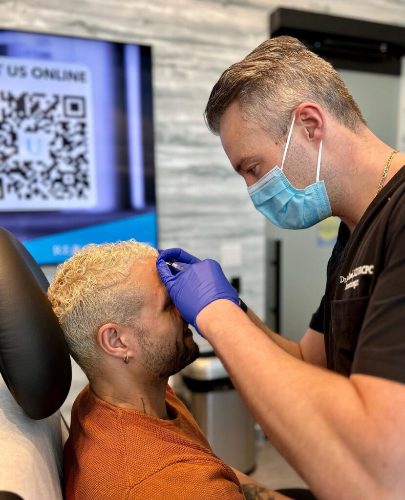 A man with curly blond hair is receiving a Botox® injection on his forehead from a healthcare professional wearing a mask, gloves, and a black uniform. The patient is seated in a clinic setting, and the healthcare professional is carefully administering the treatment with a focused expression.