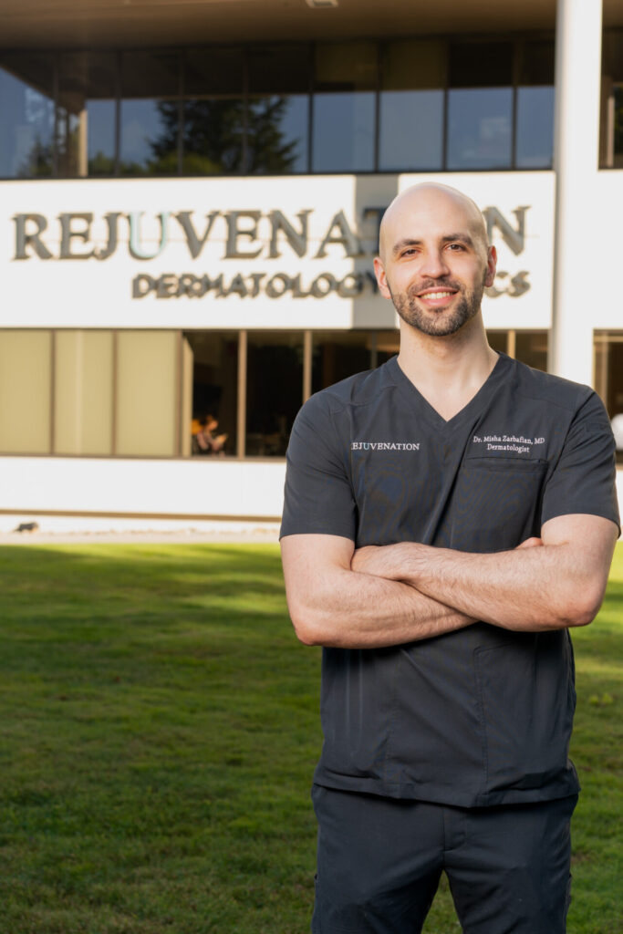 Dr. Misha Zarbafian stands confidently in front of the Rejuvenation Dermatology clinic, wearing his professional attire with a welcoming smile. The clinic's sign is visible in the background, highlighting the reputable establishment where he practices.